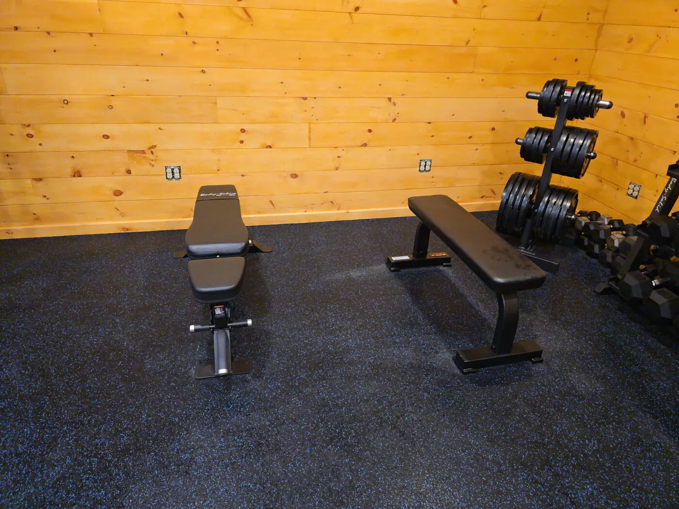 Adjustable bench, flat bench, and a plate rack on rubber gym flooring in a pine-walled home gym
